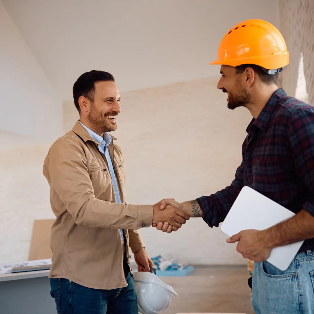 Two men shaking hands, one holding paperwork and wearing a hard hat.